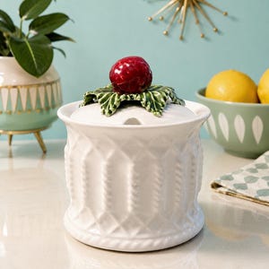 Peut inclure: Un sucrier en céramique blanche avec un motif texturé en losanges et cordes sur l'extérieur. Le couvercle présente un design sculpté de feuilles vertes surmonté d'une poignée brillante en forme de baie rouge, ajoutant un accent décoratif.