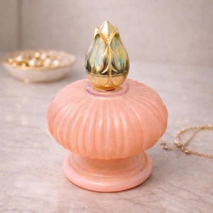 May include: A vintage-style, peach-colored perfume bottle with a ribbed body and a gold-colored, tulip-shaped stopper. The bottle sits on a marble surface. A small dish with pearls and a gold necklace are in the background.