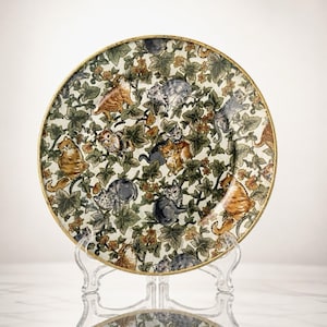 May include: A round ceramic plate featuring a detailed pattern of various cats in shades of orange, gray, and black, interspersed among green ivy leaves and small yellow flowers. The plate has a thin gold-colored rim and rests on a clear stand.