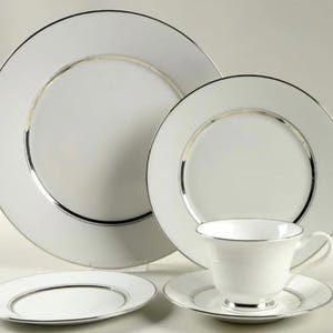 Puede incluir: Un juego de vajilla blanco con ribete plateado. El juego incluye un plato grande, un plato más pequeño, un platillo, una taza de té y un plato de pan. Los platos son redondos y tienen un diseño sencillo y elegante.