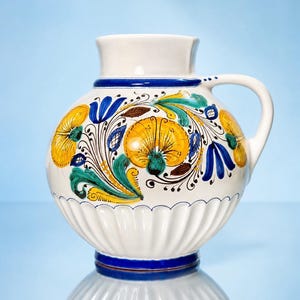 May include: A white ceramic pitcher with a rounded body and a single handle. The surface features hand-painted floral motifs in yellow, blue, and green, with a fluted base design and a solid blue band encircling the neck and the bottom edge.