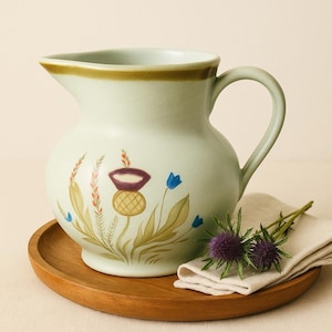 May include: A light green ceramic pitcher with a handle, decorated with floral thistle designs in purple, blue, and yellow. The pitcher sits on a round wooden tray, with a folded white cloth and purple thistles.