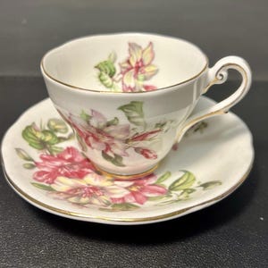 May include: A white teacup and saucer set with a floral design in pink, red, and green. The cup has a curved handle and a gold rim. The saucer also has a gold rim. The set is likely vintage or antique.