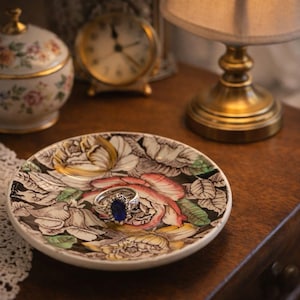 May include: A decorative ring dish with a floral design holds a silver ring with a blue gemstone. A pearl necklace and a small floral-patterned jar are nearby. A vintage clock and lamp are also visible.