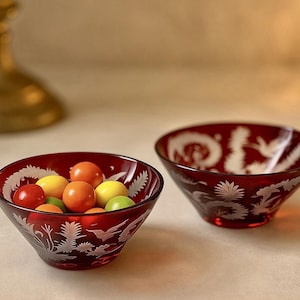 May include: Two ruby red glass bowls with etched white floral and bird designs. One bowl is filled with colorful candies. The bowls are likely decorative or for serving small portions.