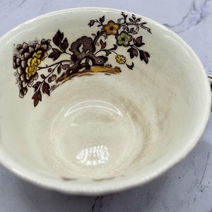 May include: Cream-colored teacup with a brown and yellow floral design. The cup has a handle and a decorative rim. The floral pattern includes clusters of yellow flowers and brown leaves. The interior of the cup shows signs of use.