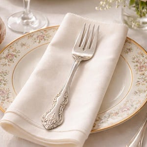 May include: A silver fork with an ornate handle rests on a folded white linen napkin. The napkin is placed on a plate with a floral design and a gold rim. A glass and a vase of pink roses are in the background, creating a formal table setting.