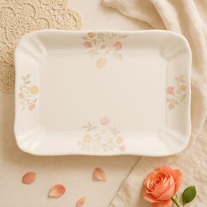 May include: A rectangular, off-white serving platter with a floral design in pink and yellow. The edges are gently scalloped. A rose and petals are arranged near the platter.