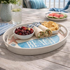 May include: An oval white serving tray with cutout handles featuring a blue graphic with the word BEACH, a starfish, and seashells. The tray holds a white bowl of blackberries, two tortilla wraps, and a small bowl of tortilla chips.