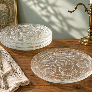 May include: A set of clear glass plates featuring a raised floral pattern of blooming flowers and leaves. The plates are round with a smooth centre and a textured rim. The design is consistent across all plates, showing clear glass construction.