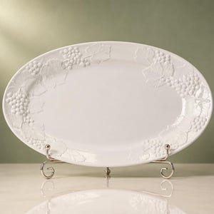 May include: An oval white ceramic serving platter featuring a raised, embossed relief pattern of grape clusters and vine leaves along the rim. The platter is supported by a silver-toned metal display stand with curved, scroll-like feet.