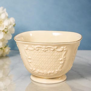 May include: A cream-coloured ceramic bowl featuring a raised lattice pattern on the exterior, framed by ornate scrollwork. The bowl has a wide, open top with a thin gold-toned rim and sits on a circular pedestal base, showcasing a glossy finish.
