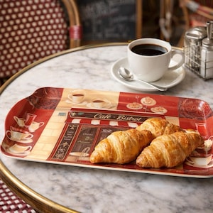 May include: A rectangular serving tray with a cafe scene design, holding three golden-brown croissants. A white coffee cup and saucer with a spoon sit nearby. The tray features a red and beige color scheme with the words "Cafe-Bistrot".