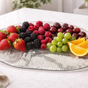 May include: A clear, oval-shaped glass platter filled with an assortment of fresh fruit. Strawberries, blackberries, raspberries, red and green grapes, and orange slices are arranged on the platter. The platter has a decorative etched design.