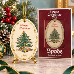 Puede incluir: Un adorno ovalado de cerámica Spode Christmas Tree con un árbol decorado, regalos, el texto Happy Holidays y el año 1996. Cuelga de un cordón dorado sobre un soporte dorado junto a su caja original de color granate con texto en blanco.