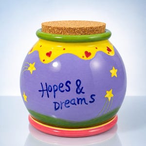 May include: A round ceramic jar with a cork lid, featuring a purple body painted with yellow stars and red hearts. The text "Hopes & Dreams" is written in blue on the front. The jar has a green rim and a pink base, with a glossy finish throughout.