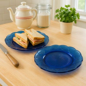 Può includere: Due piatti di vetro blu con bordo smerlato, uno con due metà di un panino. Una teiera bianca con strisce rosse, un barattolo di zucchero e una piccola pianta in vaso sono sullo sfondo. Un coltello con manico in legno è a sinistra.