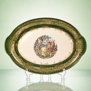 May include: An oval serving platter with a cream-colored center featuring a colorful illustration of two figures in historical attire. The wide border is dark green with an intricate gold filigree pattern throughout the entire rim.