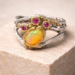 May include: A silver ring with a gold setting, featuring an oval opal gemstone and three smaller pink gemstones. The ring has a unique, textured band design, and is set on a neutral-toned surface.
