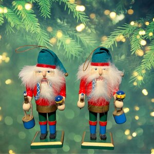 Puede incluir: Dos cascanueces navideños con chaquetas rojas, detalles verdes y azules y barbas blancas. Cada uno sostiene un adorno azul y dorado y un cubo azul. Se alzan sobre bases verdes y doradas, con un fondo borroso de ramas de árbol de Navidad.