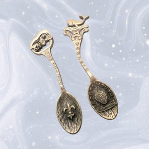Puede incluir: Dos cucharas de recuerdo metálicas con acabados antiguos. La cuchara izquierda presenta un diseño de carruaje de Nueva Orleans y un emblema de flor de lis. La derecha muestra una ballena, el texto Portsmouth New Hampshire y un icono de fresa.