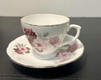 May include: A white teacup and saucer with a floral pattern featuring pink and burgundy roses. The cup and saucer have a gold trim.