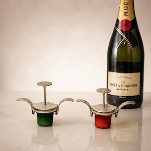 Può includere: Due tappi per bottiglie di champagne in metallo con leve laterali incernierate e meccanismo a stantuffo centrale, uno con base verde e l'altro con base rossa, posizionati accanto a una bottiglia di vetro verde scuro di champagne Moët & Chandon.