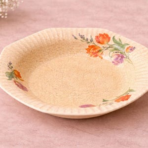 Puede incluir: Un plato de cerámica de color crema, de forma hexagonal, con una superficie texturizada. El plato está decorado con diseños florales en naranja, morado y verde. Es una pieza decorativa para servir alimentos.