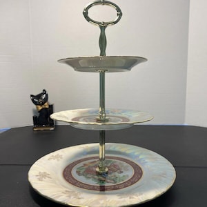 Puede incluir: Un soporte de servicio de tres niveles con un asa dorada y platos de porcelana blanca. Los platos tienen un diseño floral con detalles dorados.