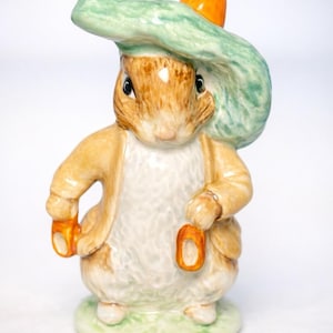 Puede incluir: Figura de cerámica de un conejo con un sombrero verde con un detalle naranja. El conejo viste una chaqueta beige y sostiene dos objetos naranjas. La figura está sobre una base verde.