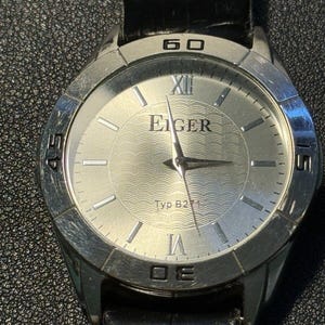 May include: A silver-toned wristwatch with a black leather band. The watch face is white with black Roman numeral hour markers and a silver-toned Eiger logo. The watch face also has a silver-toned minute hand and hour hand.