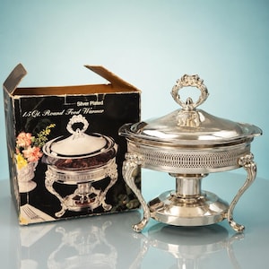 May include: A silver-plated 1.5 Qt. round food warmer with a decorative lid handle, intricate lattice side detailing, and three curved legs. The item is displayed next to its original cardboard box, which features the text Silver Plated 1.5 Qt. Round Food Warmer.