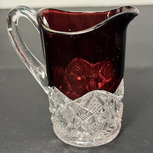 Pode incluir: Um pequeno cremeira de vidro vermelho rubi com base e alça de corte de diamante transparente. A cremeira tem um bico e uma alça transparente. A base tem um padrão texturizado detalhado. Provavelmente vintage ou antigo.