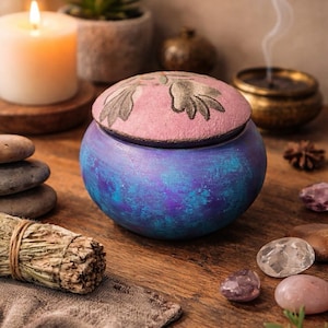 May include: A round, blue and purple decorative jar with a pink lid featuring a floral design. The jar sits on a wooden surface, surrounded by candles, stones, and a bundle of sage. The scene evokes a sense of tranquility.