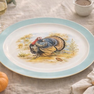 May include: A large, oval serving platter with a light blue border and a turkey illustration. The plate is surrounded by a Thanksgiving table setting, including a white pumpkin, an orange pumpkin, and a glass of wine.