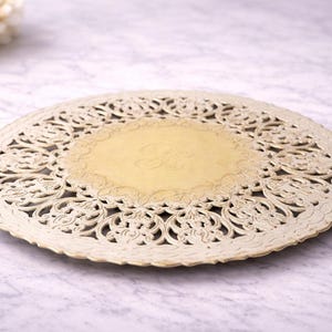 May include: A cream-colored, round doily with an intricate, openwork design. The center features a lighter, circular area with an embossed letter. Decorative floral elements are visible in the background.