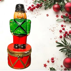 May include: A decorative Christmas ornament shaped like a toy soldier. The soldier wears a black hat, green jacket, red pants, and black boots. The ornament is standing on a red drum with gold accents. The background includes Christmas decorations.