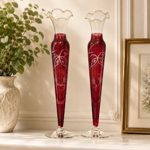 May include: A pair of tall, slender bud vases crafted from deep red glass with clear glass bases and ruffled rims. Each vase features a delicate, etched floral design on the body, showcasing a tapered silhouette and a smooth, polished finish.