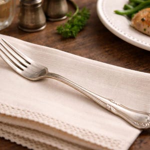 May include: A silver fork rests on a folded cream-colored linen napkin with a lace trim. The fork has an ornate handle. A plate of food and other dining items are in the background.