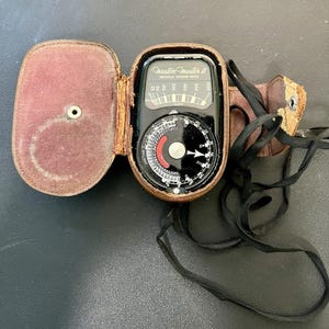 May include: A vintage Weston Master II Universal Exposure Meter in a worn leather case. The meter has a black face with white and red markings, a circular dial, and a leather strap. The case is brown with a burgundy interior.