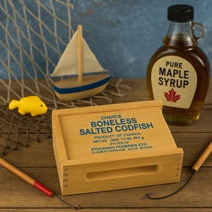 May include: A wooden box of boneless salted codfish, a bottle of pure maple syrup, a toy sailboat, and a yellow fish figurine are arranged on a wooden surface. The box has text that reads "Choice Boneless Salted Codfish".