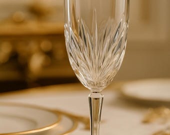Può includere: Un bicchiere da vino in cristallo trasparente con un motivo dettagliato a forma di foglia. Il bicchiere ha un gambo lungo e una base ampia. Il bicchiere è su una tovaglia bianca con piatti bordati d'oro e un coltello d'oro sullo sfondo.