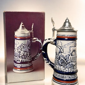 May include: A ceramic beer stein with a silver lid and handle. The stein features a blue and white design with images of wildlife and the text "Sheep-Alaskan Moose". The base has an orange and white stripe.