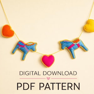 May include: A felt garland featuring two blue horses with colorful saddles and three hearts in orange, yellow, and pink, all strung on a thin yellow cord. The text DIGITAL DOWNLOAD PDF PATTERN is printed in black below the garland.