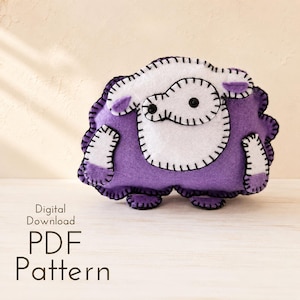 May include: A handmade felt sheep in shades of purple and white, with black stitched details. The sheep has a rounded body, small ears, and black button eyes. The text "Digital Download PDF Pattern" is below the sheep.