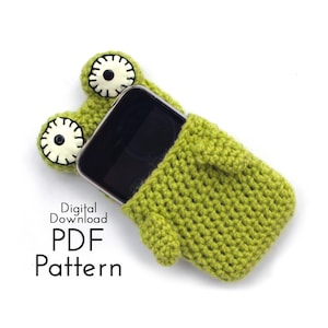 May include: A green crocheted phone case shaped like a frog, with large white and black eyes. The phone is partially visible inside the case. Text on the image reads "Digital Download PDF Pattern".