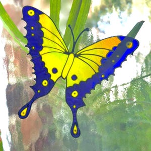 May include: A vibrant butterfly decoration featuring yellow and blue wings. The butterfly has a detailed design with yellow spots and a yellow body. The wings are outlined in black, set against a blurred background of green foliage.