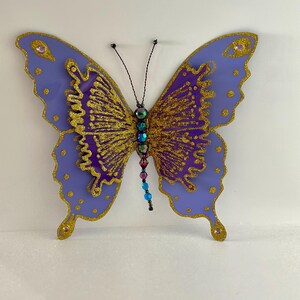 May include: A large, decorative butterfly with purple wings outlined in gold glitter. The center of the wings is filled with gold glitter. The butterfly has a beaded body with a variety of colorful beads and antennae.
