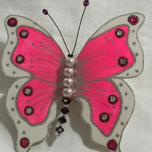 May include: A decorative butterfly with bright pink wings outlined in silver glitter. The wings have white edges with silver dots and pink and clear gem accents. The butterfly's body is made of pearls and dark purple beads, with black wire antennae.