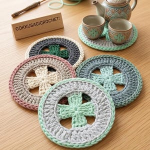 K&ouml;nnte beinhalten: Eine Sammlung handgefertigter H&auml;keluntersetzer in verschiedenen Farben, darunter Grau, Rosa und Gr&uuml;n, mit einem kreuzf&ouml;rmigen Design in der Mitte. Das Bild zeigt auch eine Teekanne, Teetassen und ein Holzschild mit der Aufschrift "GOKKUSAGICROCHET:" und dem Text "PDF CROCHET PATTERN".